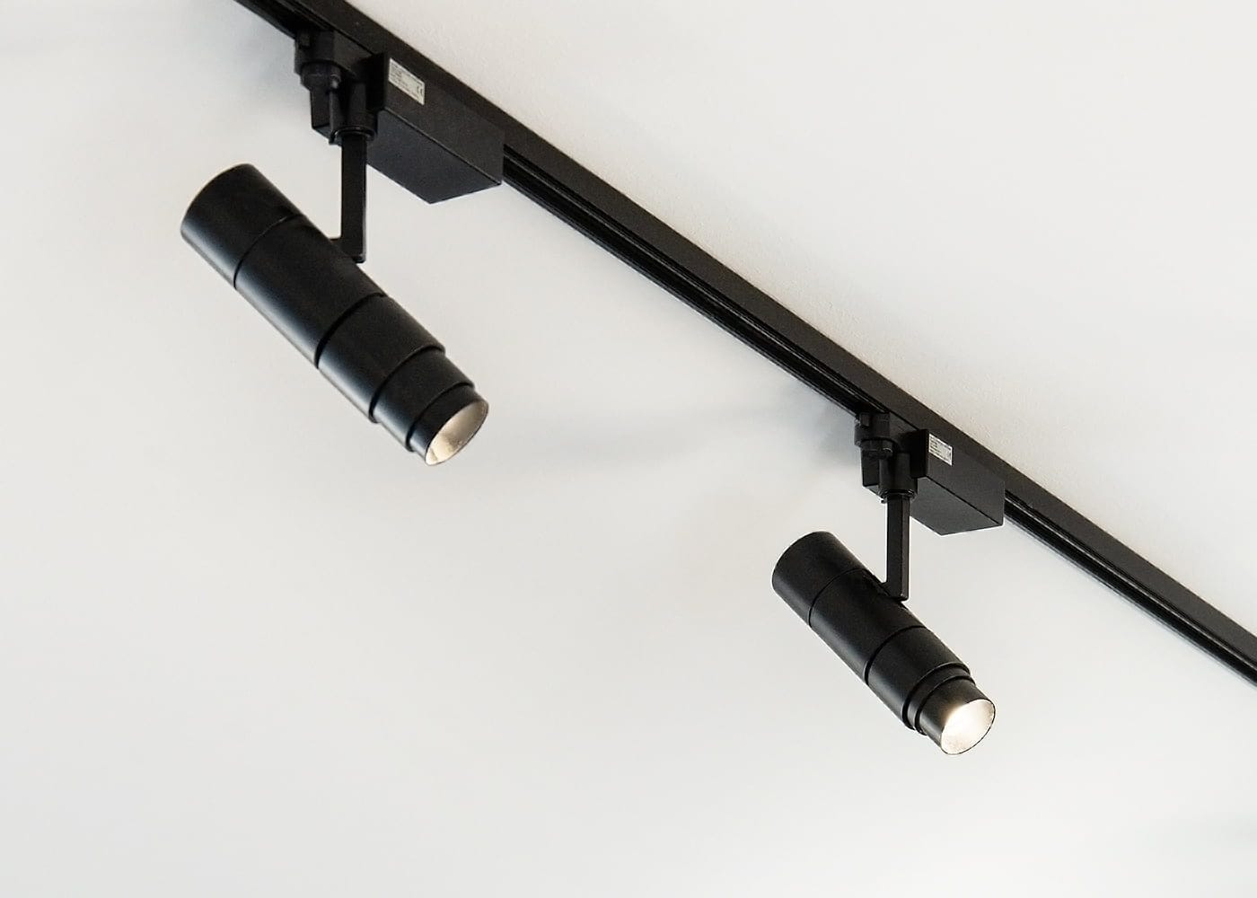 ZOOM-kiskospotti-blogi-banneri-ledstore.fi - LedStore Minimalistinen kokoonpano kahdella mustalla sylinterimäisellä LED-kiskovalolla valkoisella katolla, kiinnitettynä mustaan kiskoon, esittelee modernia valaistussuunnittelua ja säädettäviä kulmia.