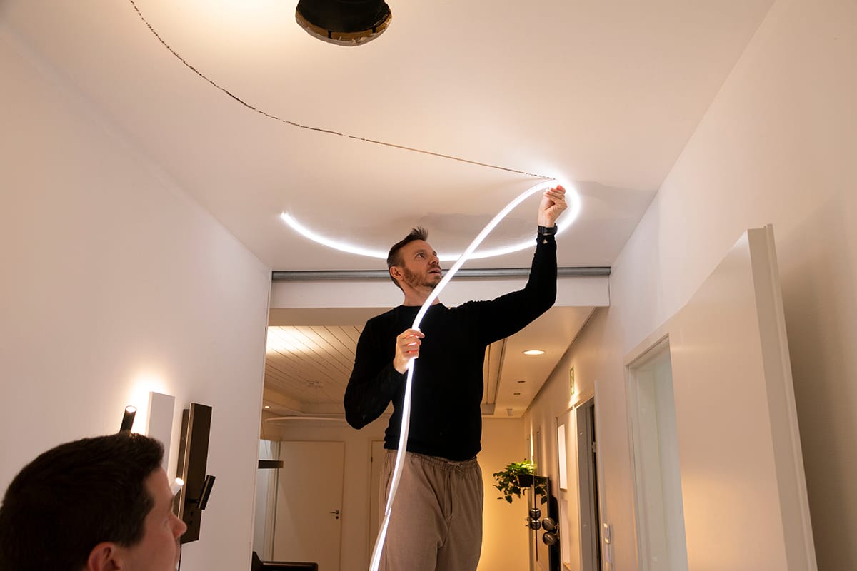 IMG_8379 kopio 2 - LedStore Henkilö kiinnittää joustavan LED-nauhavalaisimen moderniin kattoon, mikä parantaa huoneen valaistusta. Musta villapaita, beige housut; toinen henkilö etualalla, epätarkka.