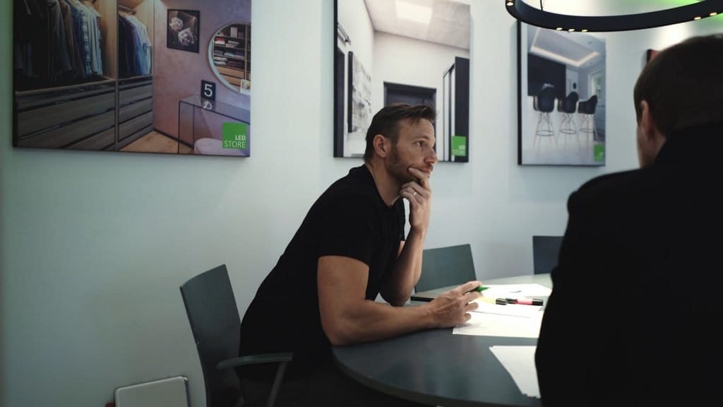 Janne suunnittelee - LedStore Mies, jolla on parta pöydässä, leuka kädellä, modernissa huoneessa; LED-valaistut julisteet seinillä esittelevät kaappimalleja. LED-valaistus myymälän tunnelmaa.