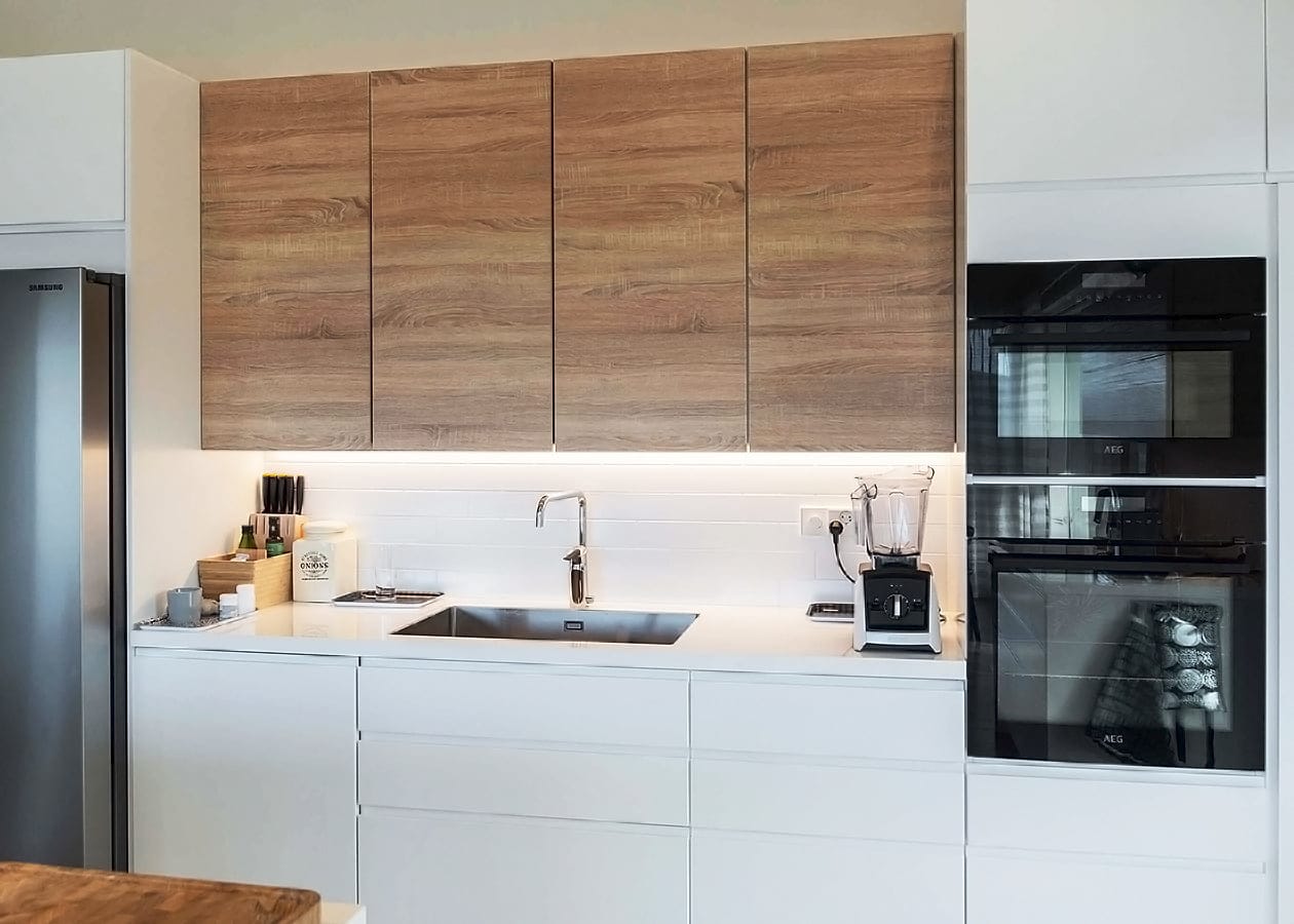 Keittiön työtasovalaistus led-nauhalla_ledstore.fi - LedStore Moderni keittiö, jossa on puiset yläkaapit, LED-valaistut valkoiset työtasot, ruostumattomasta teräksestä valmistettu pesuallas kromihanalla, kaksinkertainen uuni ja työtasolaitteet.