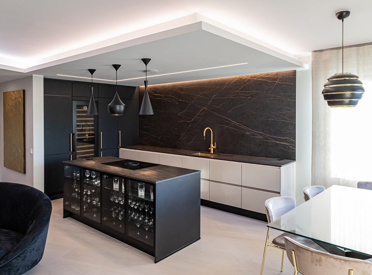 Moderni keittiö, jossa on tilaat mustat ja valkoiset ka valkoiset ka, mustasta marmorista tehty välitila ja kullanvärinen hana. Riippuvalaisimet valaisevat mustaa saareketta, jossa on vuosi lasiastioita. Vieressä sijaitsevassa lasisessa ruokapöydässä on verhoillut tuolit, ja sen yläpuolella loistaa näyttävä LED-korostettu musta-kultainen riippuvalaisin. Tutustu LedStore.fi:n laajaan LED-valaistusvalikoimaan luodaksesi vastaavan upean ilmeen omaan kotiisi!