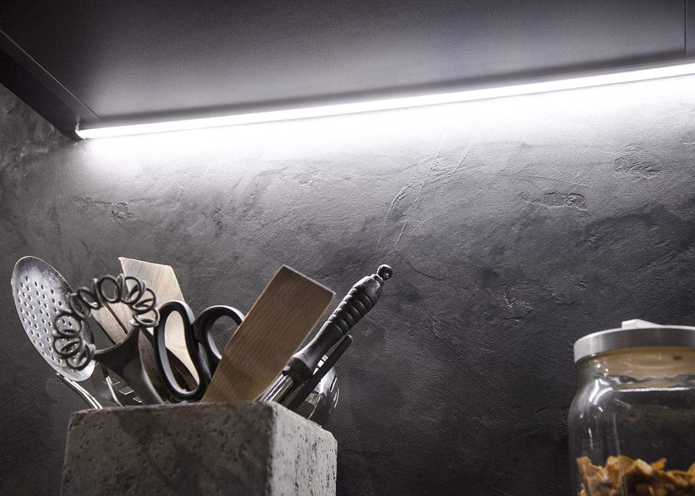 Keittiön led nauha valaistus - LedStore Keittiökuva, jossa astiat pidikkeessä, kirkas LED-nauhavalo korostaa kuvioitua tummaa seinää; lasipurkki näkyy oikealla. Ihanteellinen moderniin LED-valaistukseen.