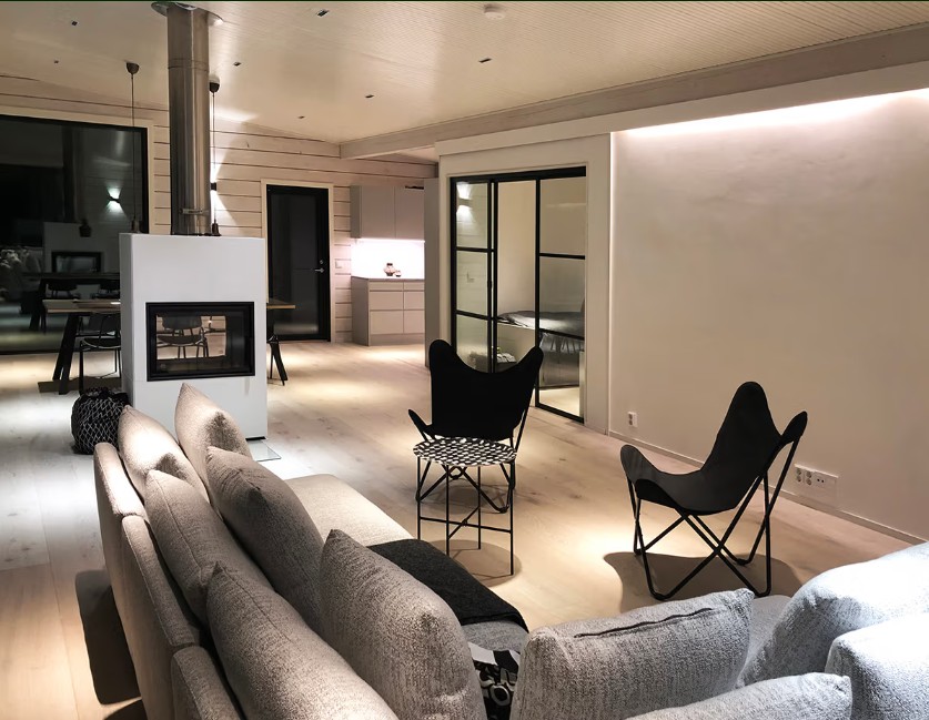 LedStoren LINJA 5 -spottivalaisimet pääroolissa mökin valaisussa - LedStore Moderni olohuone, jossa harmaa sohva, mustat tuolit, takka ja taustalla avokeittiö – tunnelman ja tyylin muotoiluee LedStore.fi:n laadukas led-valaistusratkaisu.