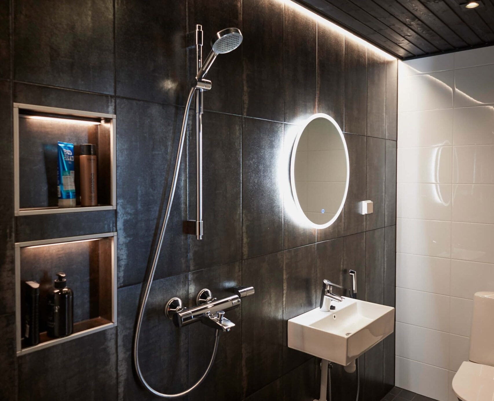 Tyylikäs kylpyhuone tummat laatat, LED-taustavalaistu peili pesualtaan yläpuolella, suihku, sisäänrakennetut hyllyt ja musta katto. Moderni wc täydentää tyylikkään ilmeen.