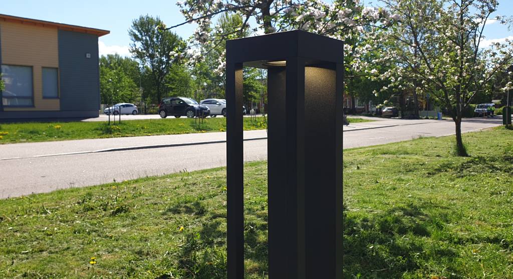 lähikuva valot päällä FUNK PILLAR - LedStore Moderni musta valaisinpylväs LED-valolla tien varrella; kukkivia puita, pysäköityjä autoja ja rakennus, jossa on suuret ikkunat taustalla aurinkoisen taivaan alla.