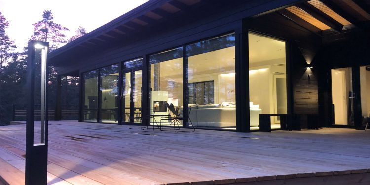 piha2-750x375 - LedStore Moderni talo, jossa on suuret lasi-ikkunat ja tilava puinen terassi, ympäröivätynä puilla. Sisätilat ovat kirkkaasti valaistut ja esittelevät minimalistisen designin käsissä sävyissä. LedStore.fi:n LED-ulkovalot lisää lämpimän hehkun iltahämärään.