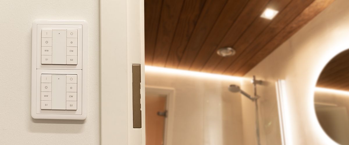 valo_zigbee_ohjaimet_1_kansikuva - LedStore Kylpyhuone, jossa on puiset kattopaneelit ja pehmeä LED-valaistus. Tilasta löytyy myös kätevä walk-in-suihku sadesuihkupäällä. Läheisellä seinällä on kaksi modernia valokytkimen paneelia, ohjataan näppäimillä. Tyylikkään kokonaisuuden kruunaa pyöreä led-valaistu peili.